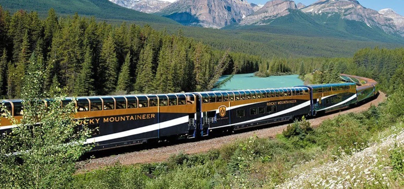 A Rocky Mountaineer train traveling through a lush green forest with a mountain range in the background.