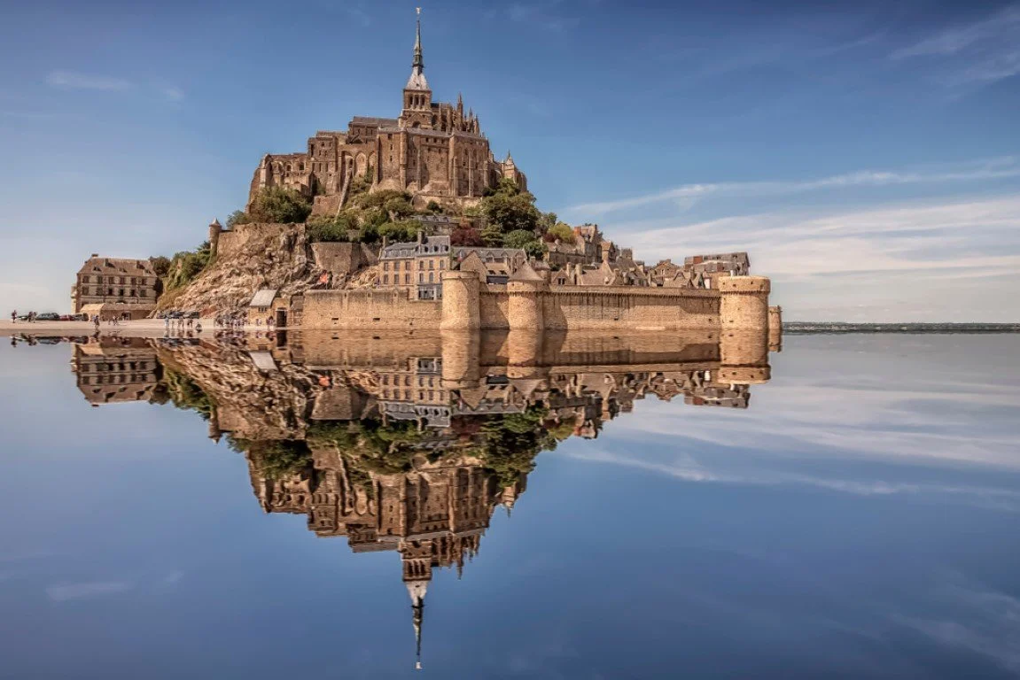 Mont Saint-Michel, a historic island commune with a medieval monastery, reflected in the surrounding water under a blue sky.