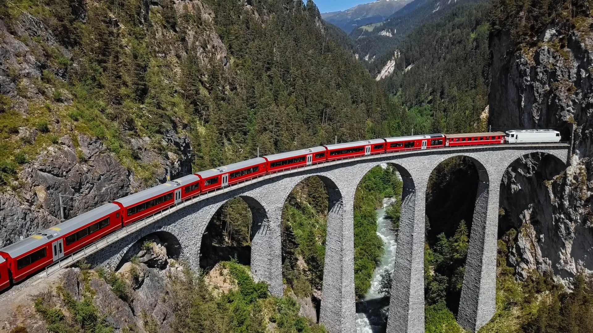 A red train traveling across a stone arch bridge over a river in a mountain canyon with dense evergreen trees on the slopes.