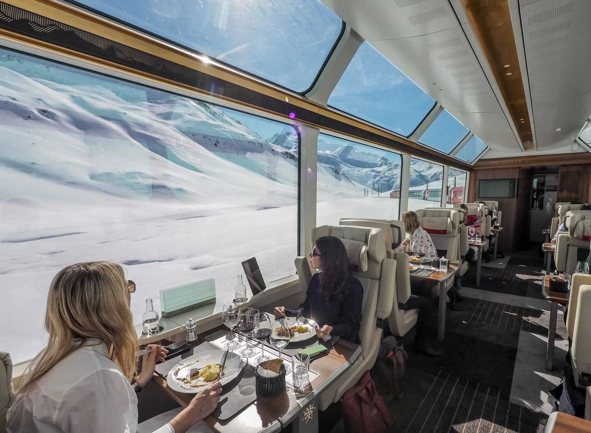 People dining in a train restaurant with large windows showing snowy mountains and a train track outside.
