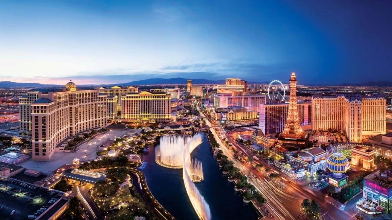 Nighttime aerial view of Las Vegas Strip with illuminated hotels, casinos, and landmarks including the Eiffel Tower replica, a large Ferris wheel, and the Bellagio fountain show, with colorful lights and a dark sky.