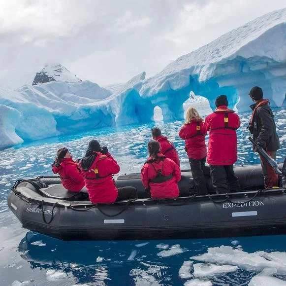 Group of people in red jackets in an inflatable boat near icy blue glaciers and icebergs in an Arctic or Antarctic landscape.
