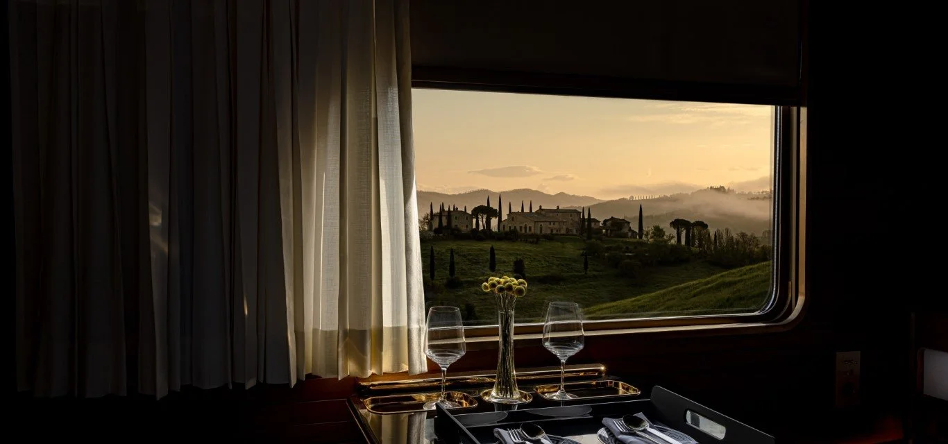A table set with two wine glasses, a vase with flowers, and silverware by a window overlooking a scenic landscape with houses, trees, hills, and fog at sunset.