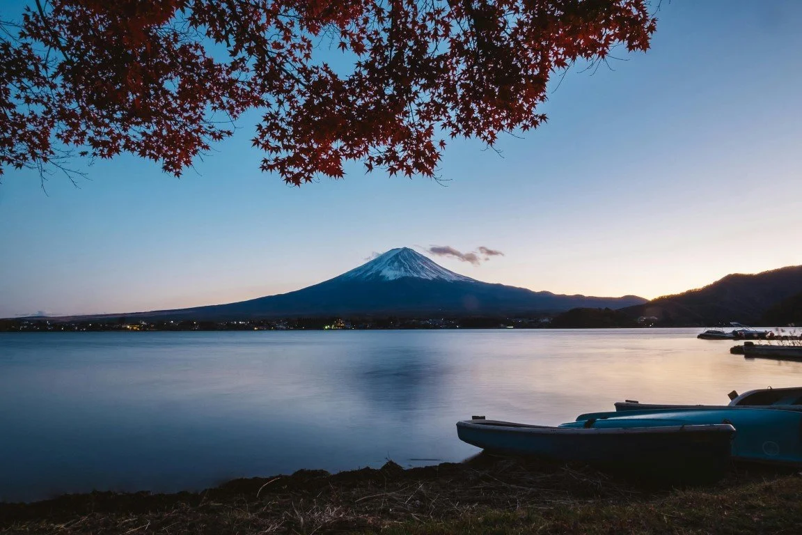 Mount Fuji overlooking a lake at sunset, with boats on the shore and autumn leaves in the foreground.
