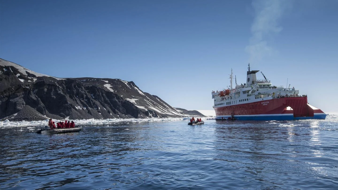 An expedition ship navigating icy waters near a snowy mountain coastline, with several smaller boats carrying people on board.