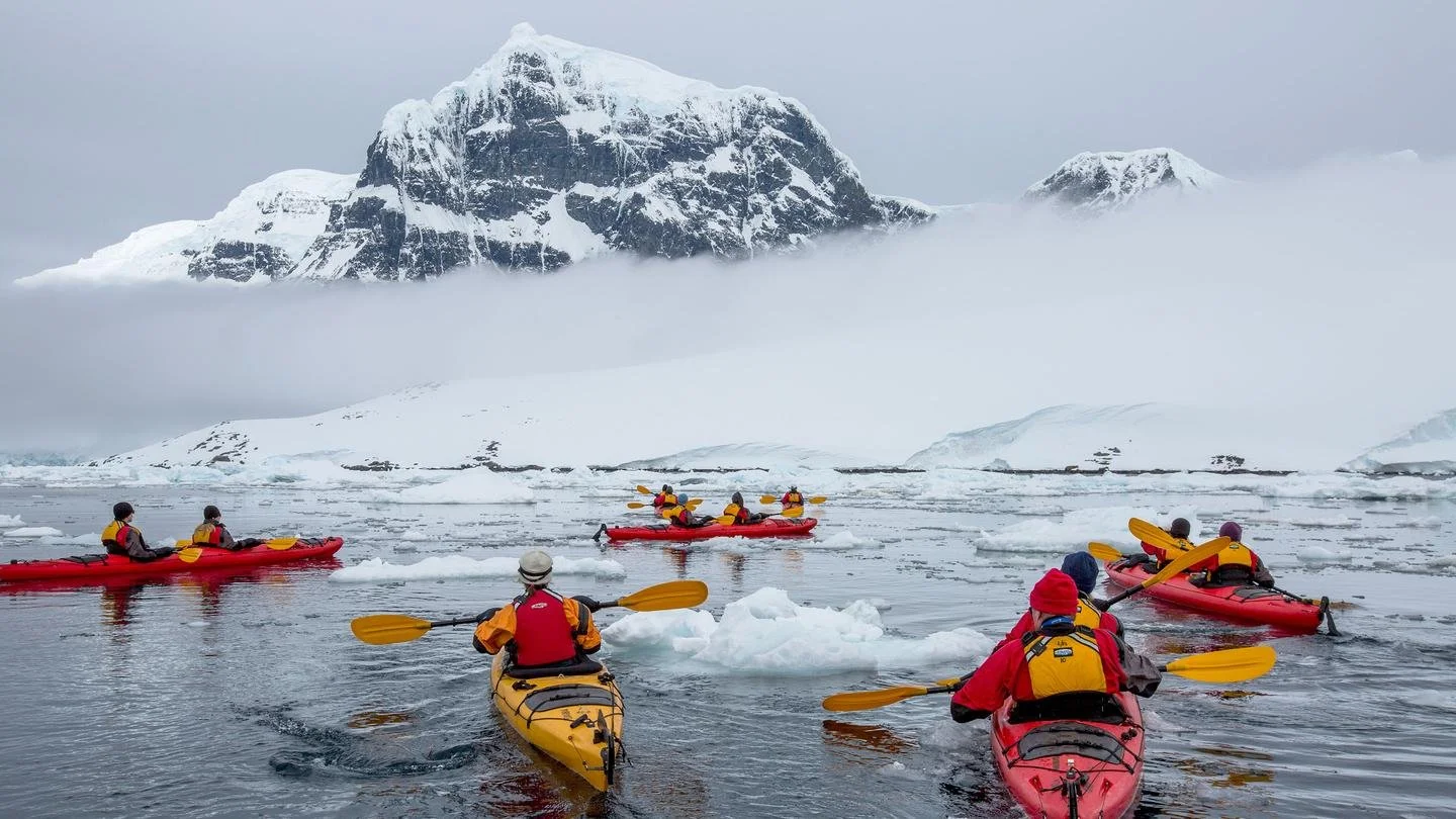 A group of people kayaking in icy waters near snow-capped mountains with fog and icebergs around.