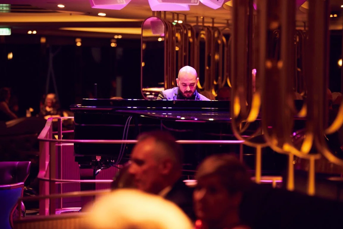 A man playing a grand piano on a dimly lit stage with purple and orange lighting at a live music venue.