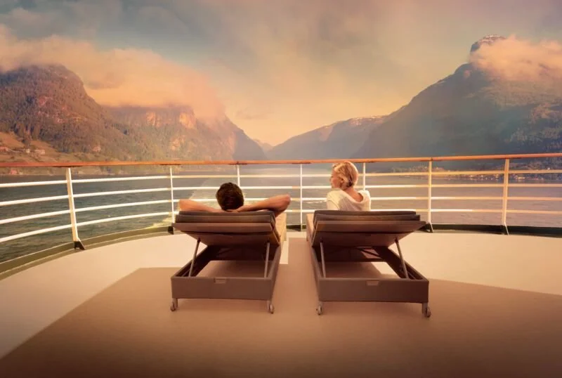 Two people relaxing on lounge chairs on a ship's deck, looking out at a scenic fjord with mountains and a colorful sky at sunset.