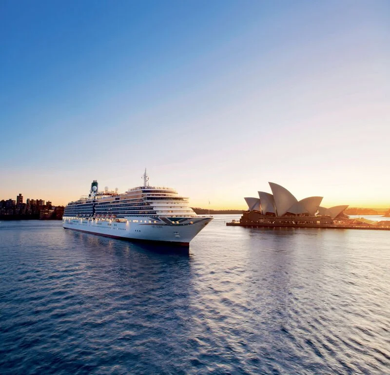 A large white cruise ship sailing near the Sydney Opera House during sunset.