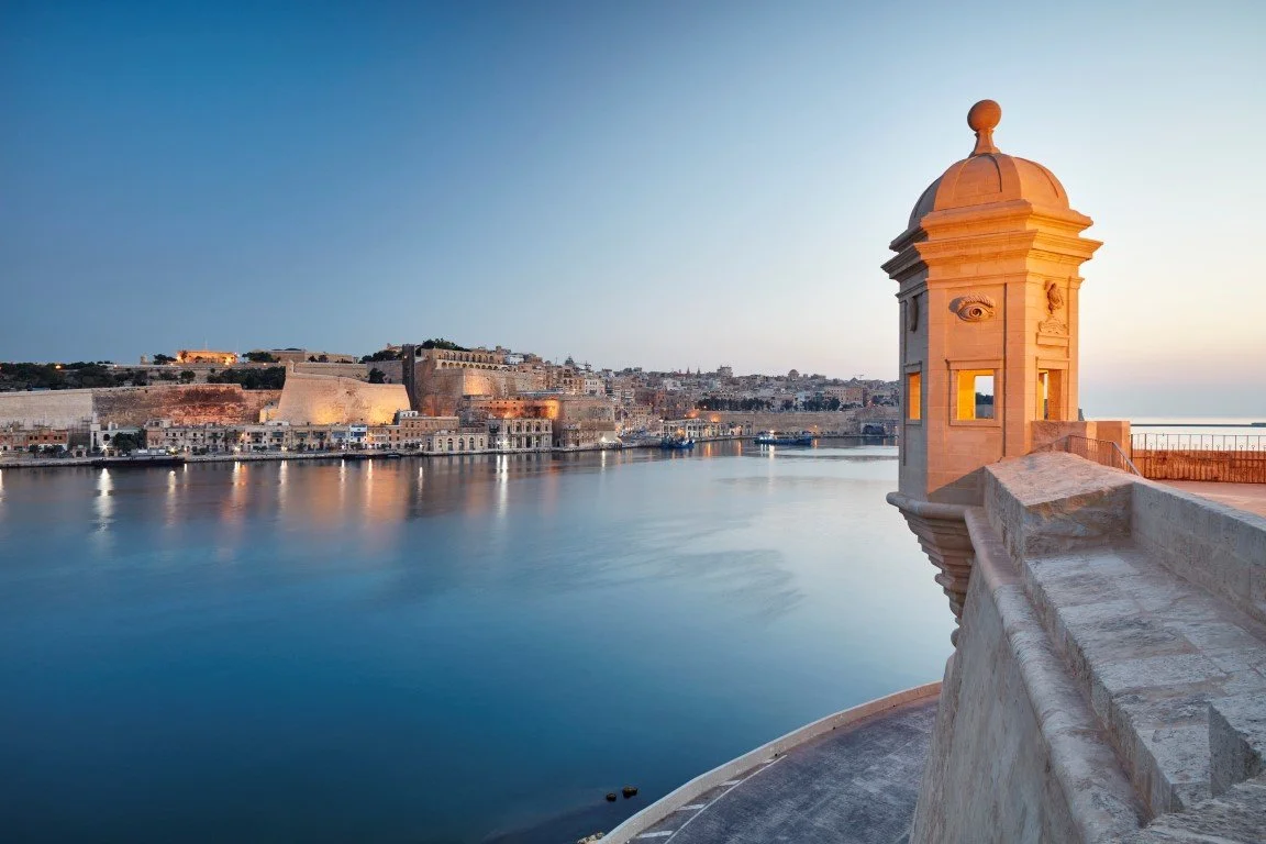 View of a calm waterfront with historic buildings on the shoreline at sunset, featuring a stone watchtower in the foreground.