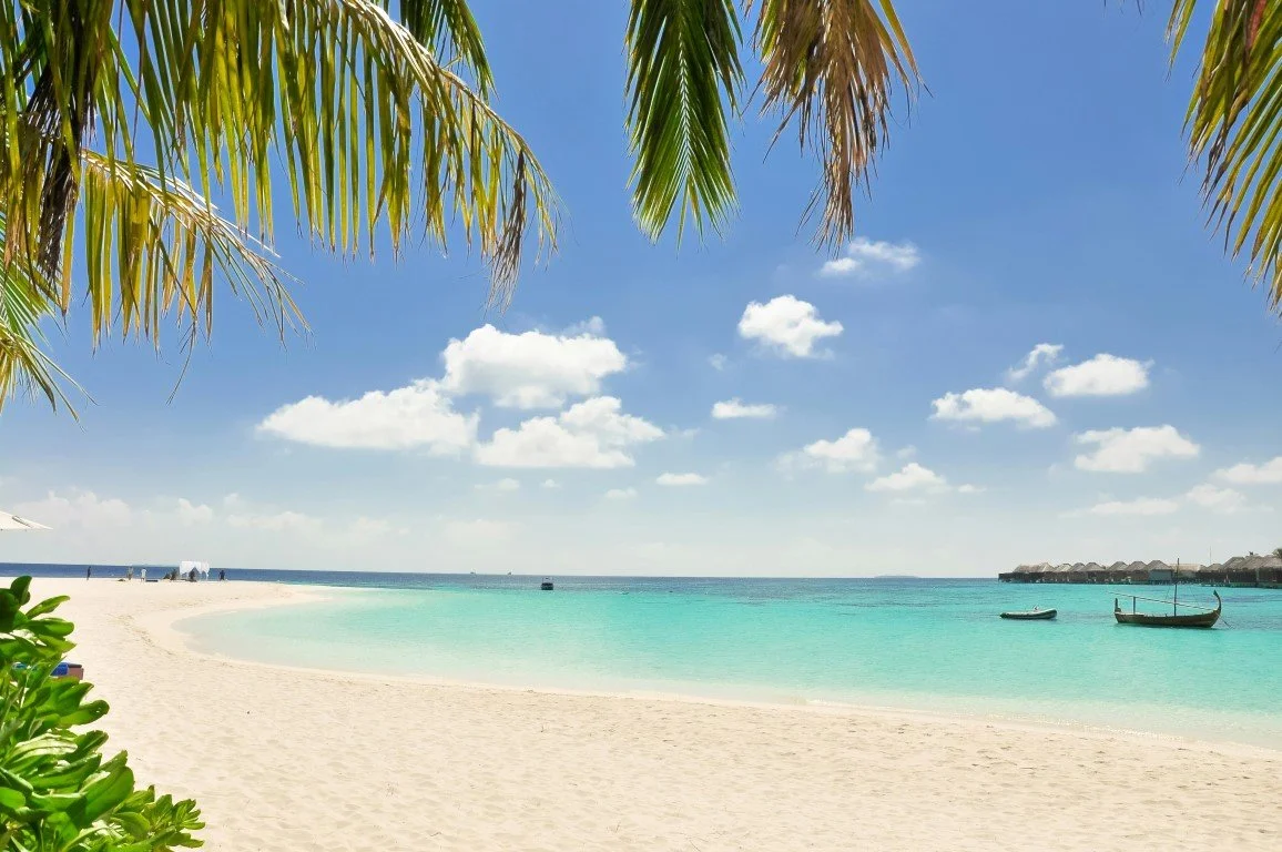 A tropical beach scene with white sand, turquoise water, and a clear blue sky with scattered clouds. Palm trees frame the top of the image, and there are a few small boats floating near the shore.