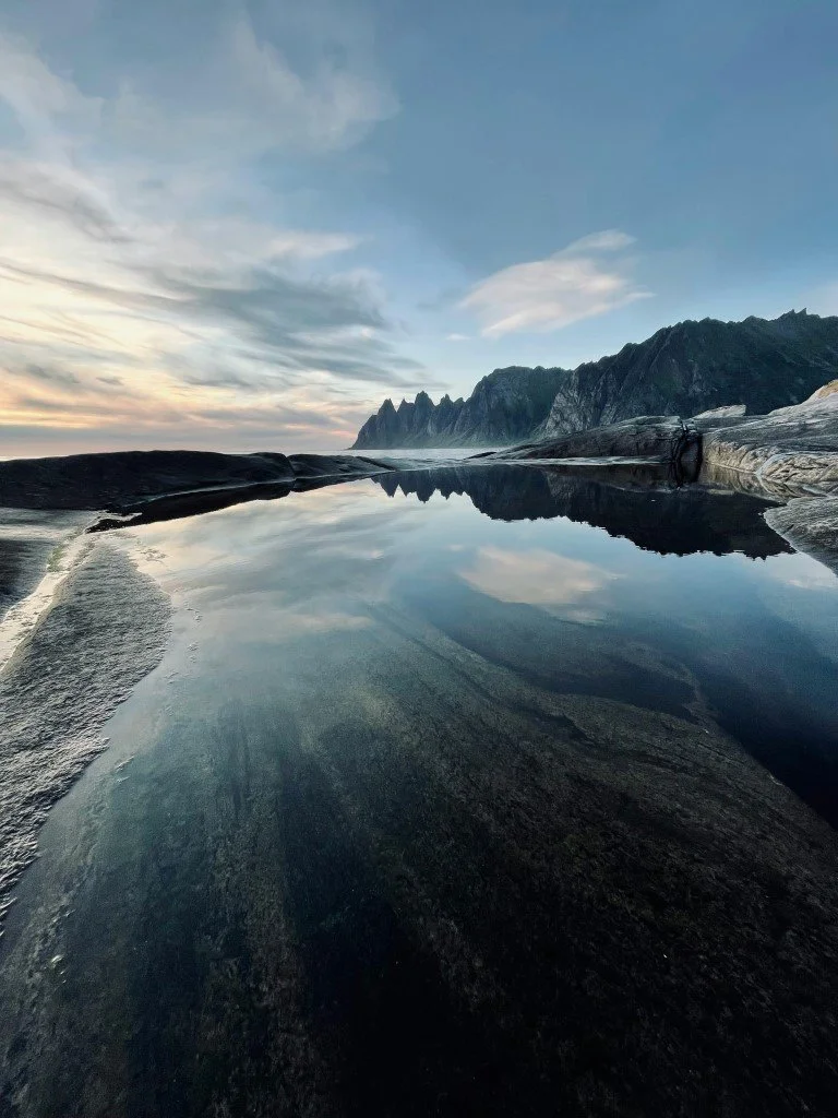 A reflective tide pool on a rocky shoreline with a mountain range in the distance and a partly cloudy sky.