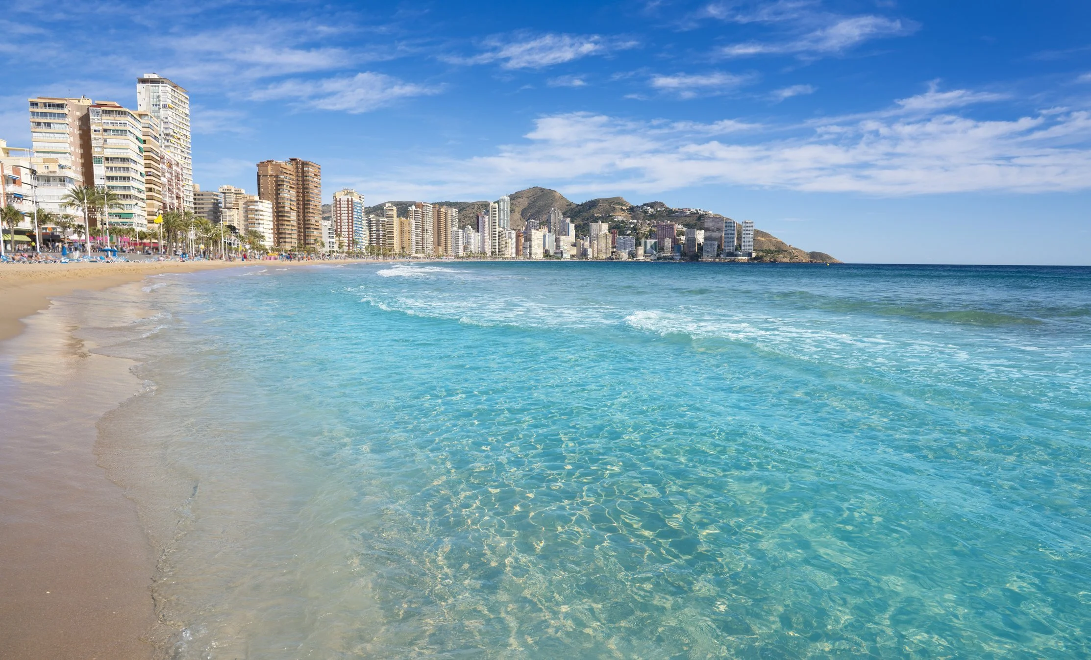 Sunny beach with clear blue water and high-rise buildings along the shoreline under a blue sky with scattered clouds.