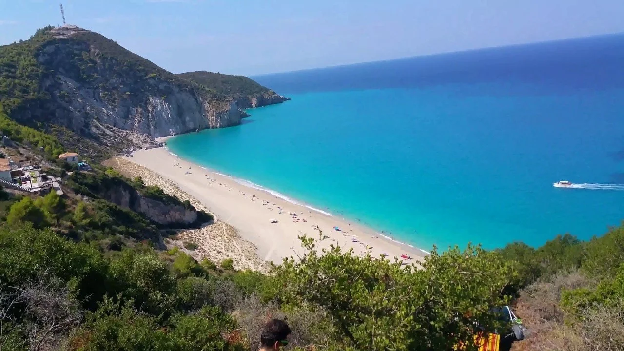 A scenic view of a beach with turquoise waters, white sand, green trees in the foreground, and cliffs in the background with a few buildings on the hillside. A boat is sailing in the water.