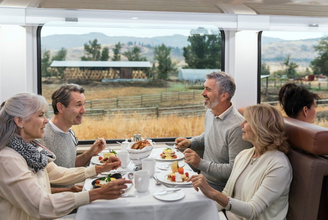 Five seniors enjoying a meal together inside a train, with a rural landscape visible through the window behind them.