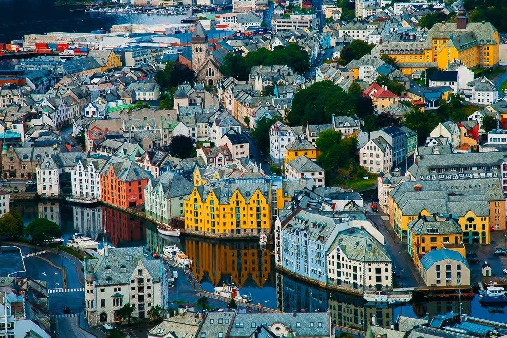 Colorful waterfront buildings and houses in a European city, with a church steeple, waterway and boats, surrounded by greenery.