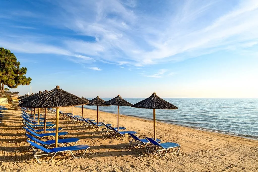 Empty beach with blue lounge chairs and thatched umbrellas, calm sea, and partly cloudy sky during daytime.
