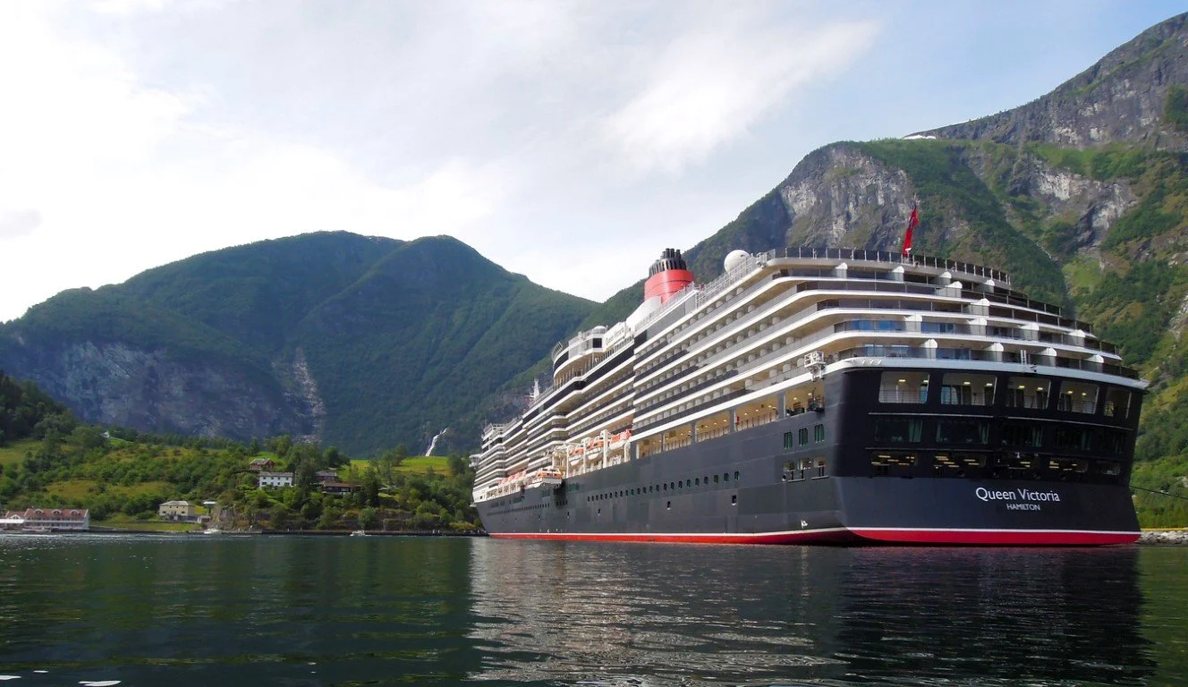 A large cruise ship named Queen Victoria near a green mountainous coastline.