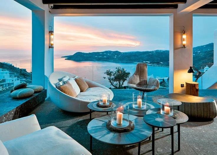 Open-air living space with white and gray furniture, candles on marble-topped tables, overlooking a coastal landscape during sunset.