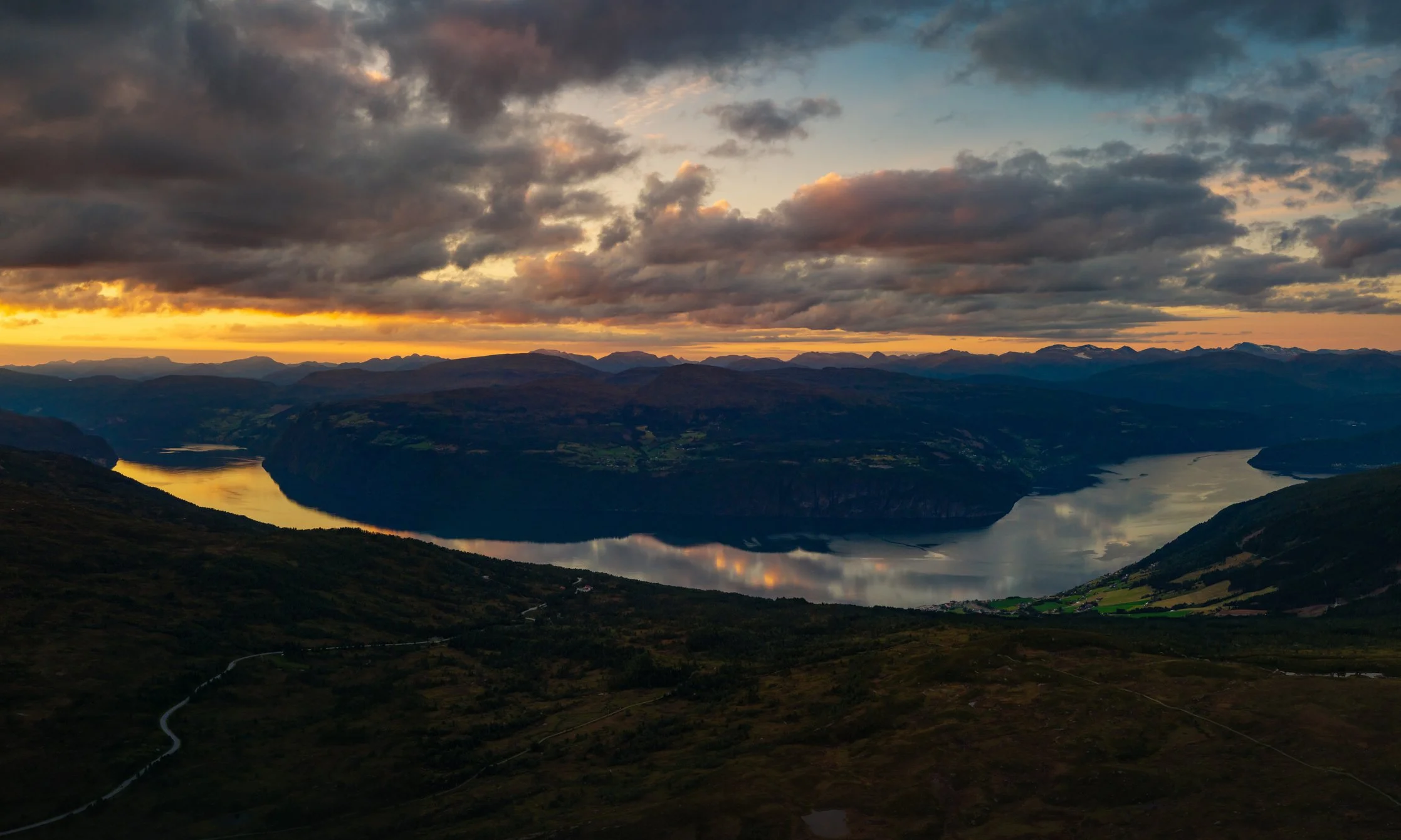 A scenic view of a winding river flowing through a valley with mountains in the background, under a dramatic cloudy sky at sunset.