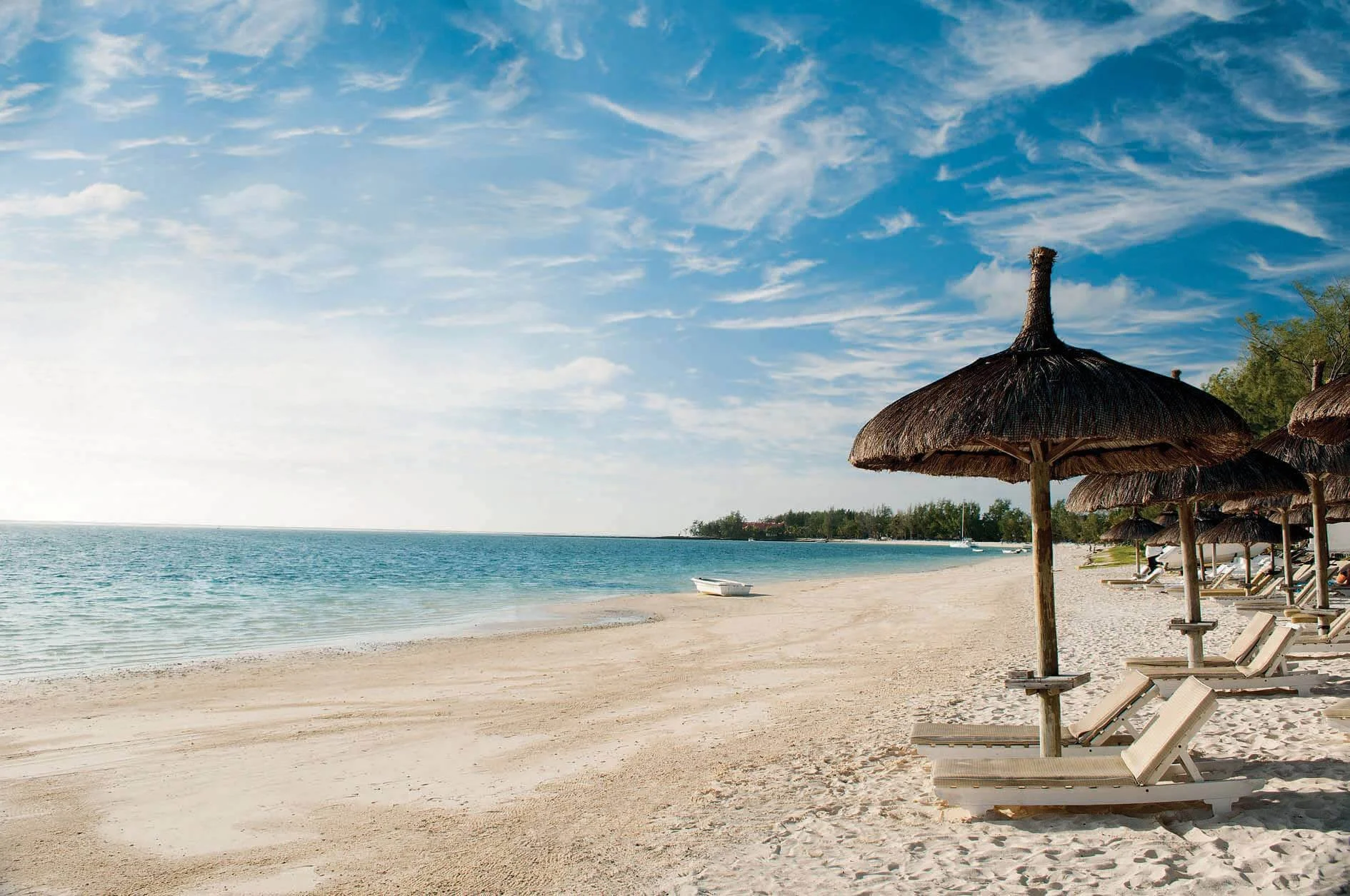 A tropical beach with white sand, blue water, and straw umbrellas with lounge chairs along the shoreline under a partly cloudy sky.