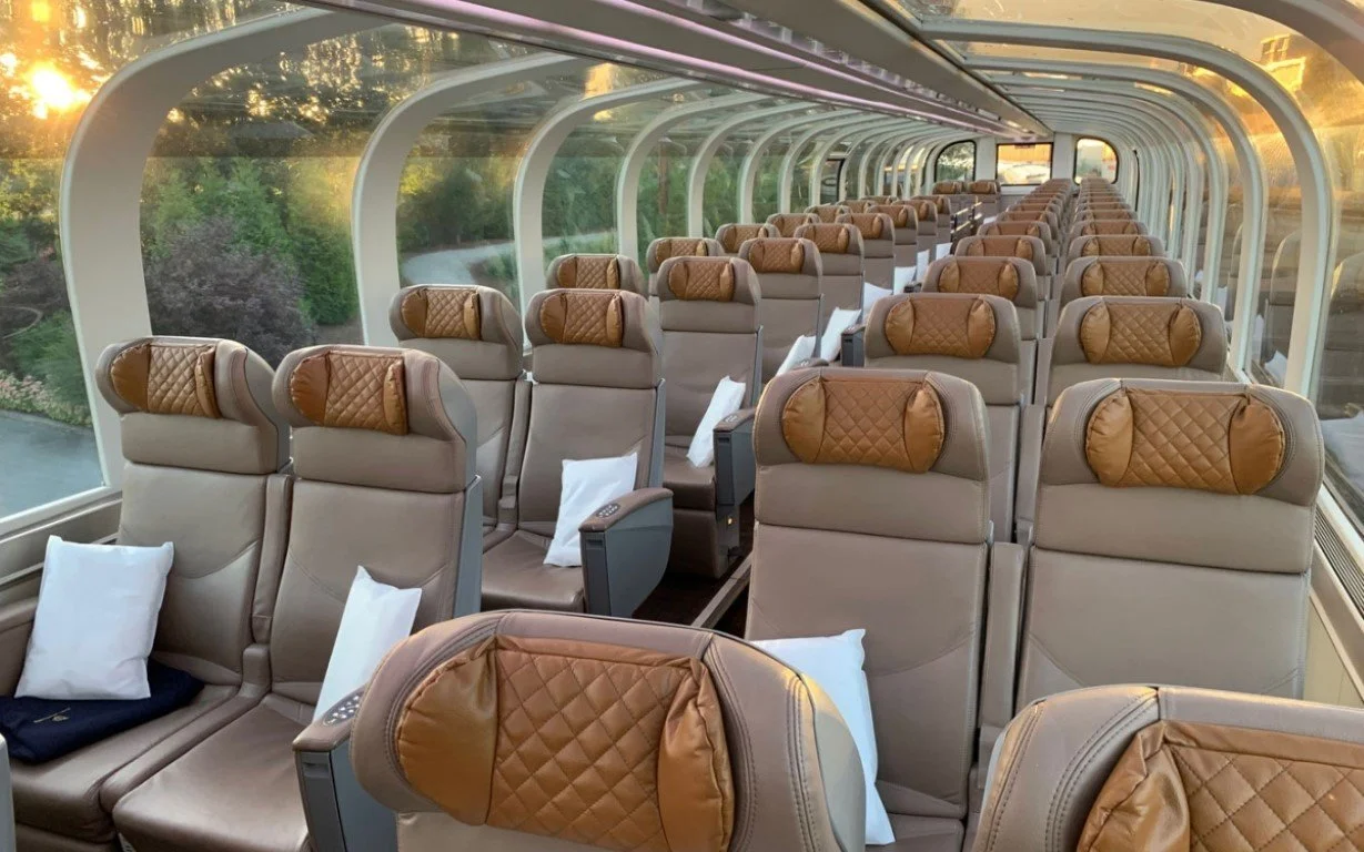 Interior of Canadian Rockies Train with multiple beige seats, each with a brown quilted headrest and white pillows, during sunset with a view of the Rockies  outside.
