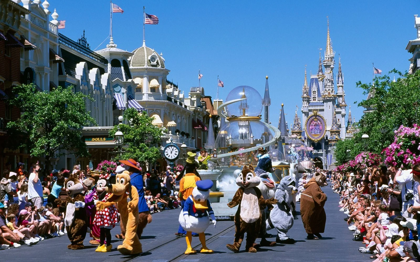 A parade featuring Disney characters walking down Main Street at Disneyland with the castle in the background, surrounded by seated spectators and fans.