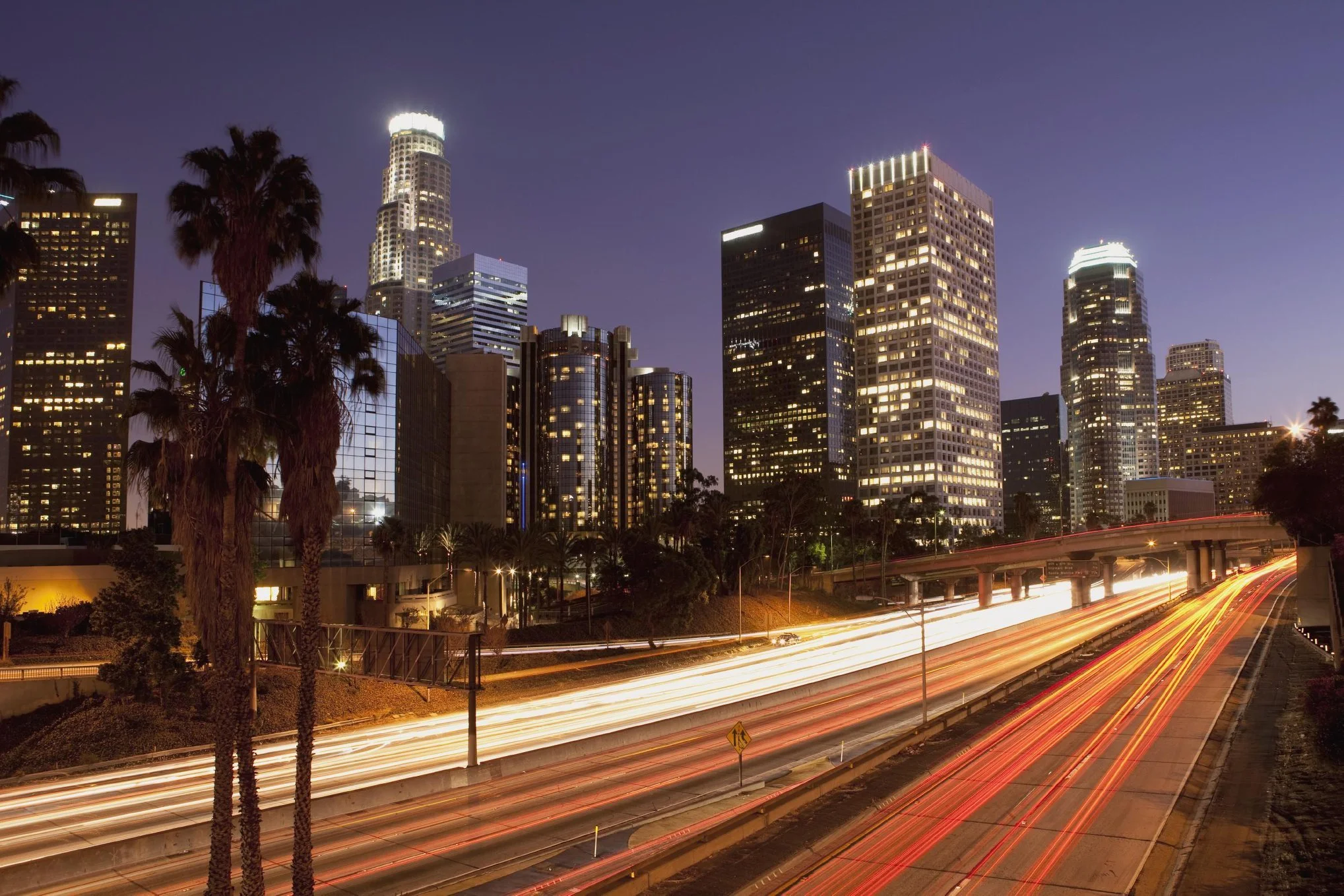 Nighttime city skyline with tall illuminated skyscrapers and streaks of car light trails on the highway in Los Angeles.
