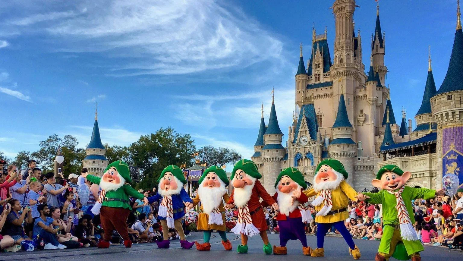 Disney parade featuring seven dwarfs dressed as Christmas elves holding hands in front of Disneyland castle with audience watching.