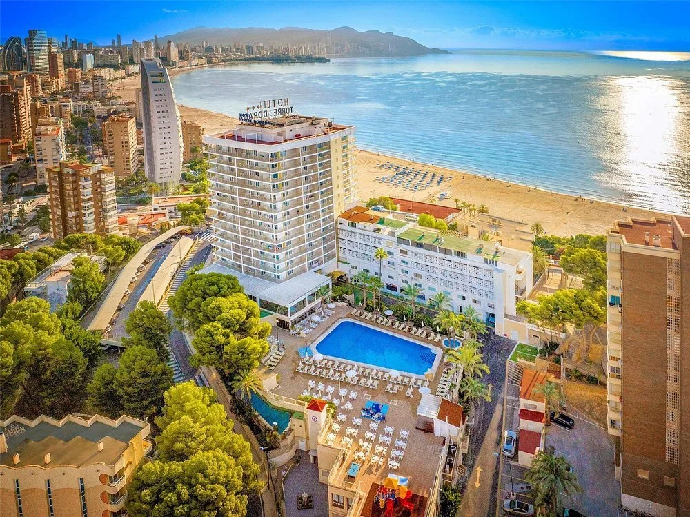 Aerial view of a beachfront hotel with a large swimming pool, surrounded by lounge chairs and palm trees, overlooking the ocean and a sandy beach in a city with tall buildings and mountains in the background.