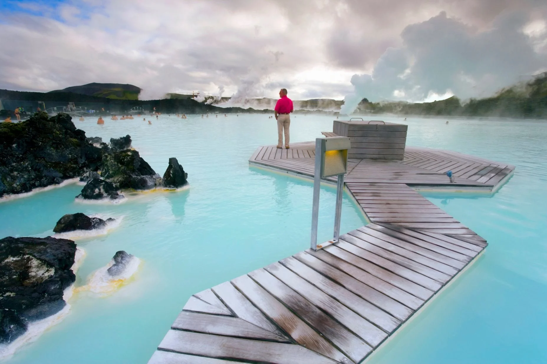 A person stands on a wooden dock extending into hot spring geothermal pools, with steaming water and volcanic rocks nearby, and cloudy skies overhead.