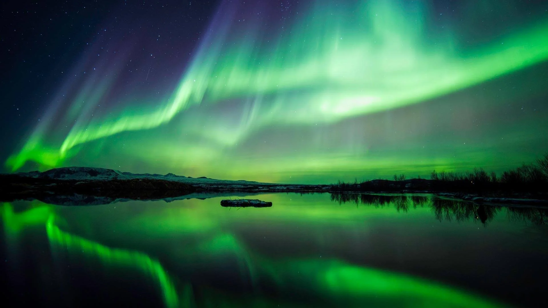 Northern lights glowing green in the night sky over a calm body of water, reflecting the aurora and silhouettes of distant land and trees.