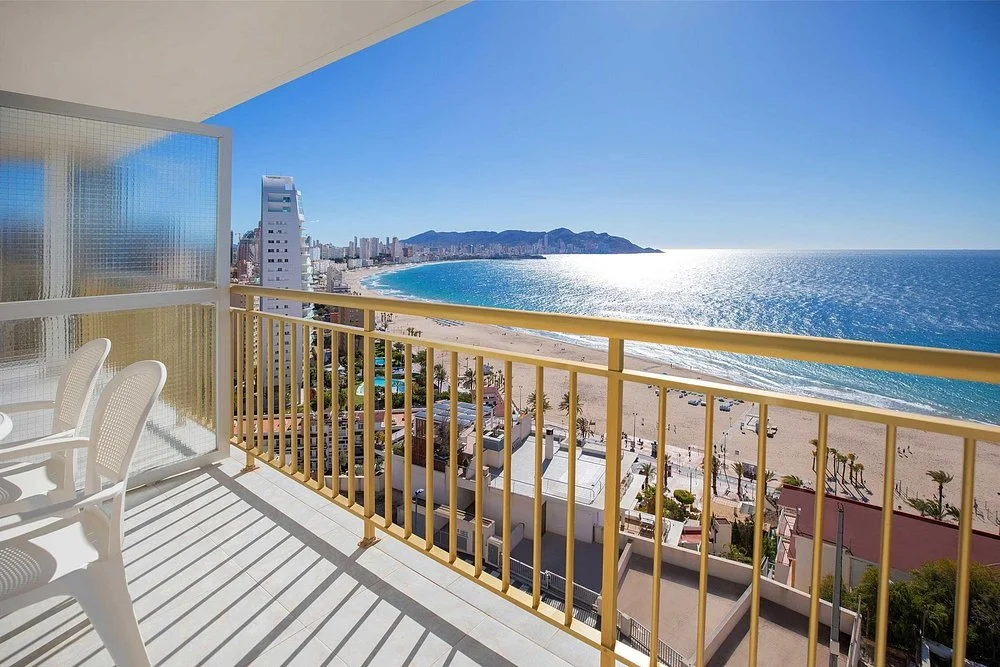 View from a balcony overlooking a beach and ocean with city skyline and mountains in the background.