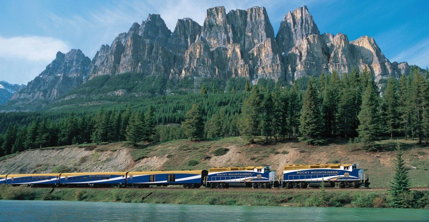 A Rocky Mountaineer train traveling along a river with a backdrop of green forest and tall mountain peaks.