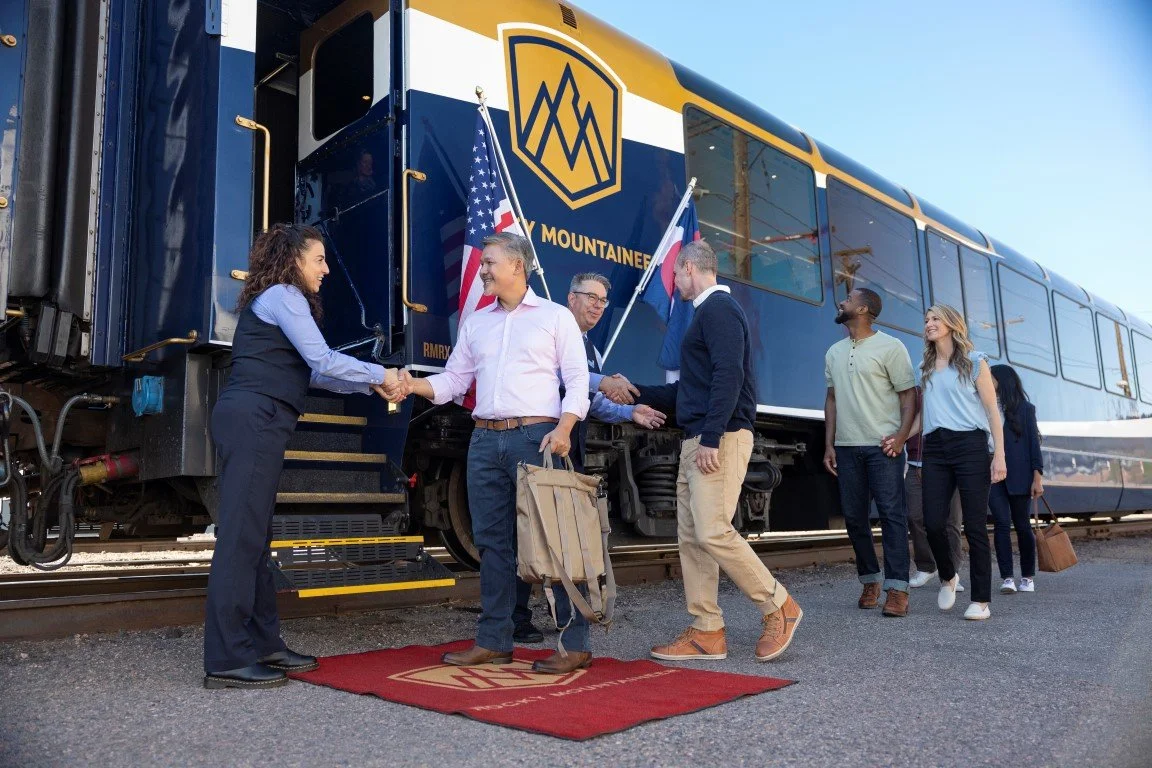 People greeting each other on a train platform in front of a the rockies train with gold accents and a mountain logo, with American flags visible in the background.