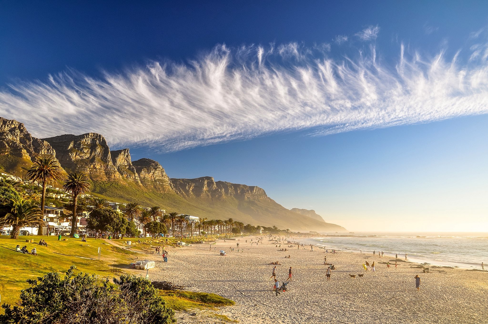 A beach scene with people walking and sitting on the sand, palm trees along the shoreline, mountain cliffs in the background, and a partly cloudy sky with wispy clouds overhead during sunset or late afternoon.