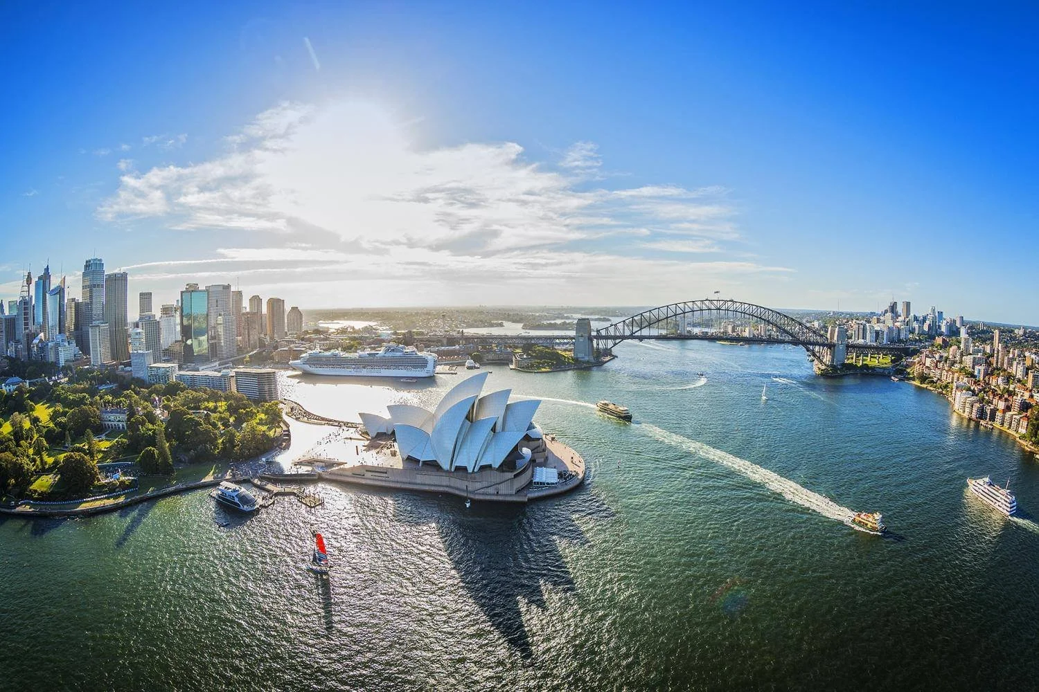 Aerial view of Sydney Harbour showing the Sydney Opera House, Harbour Bridge, city skyline, and boats on the water on a sunny day.