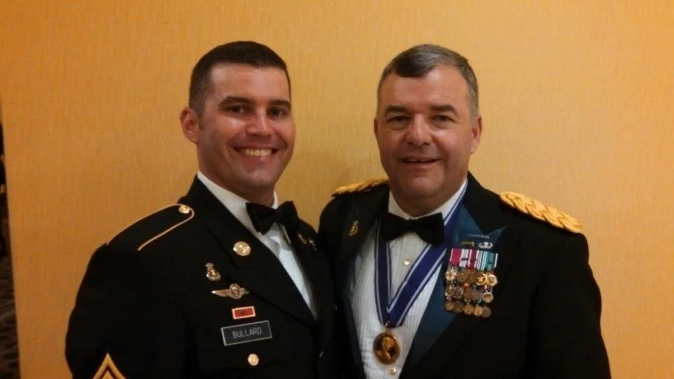 Two men in military formal uniforms, smiling, standing together against a yellow wall. One has a name tag reading 'Bullard' and the other wears numerous medals and ribbons.