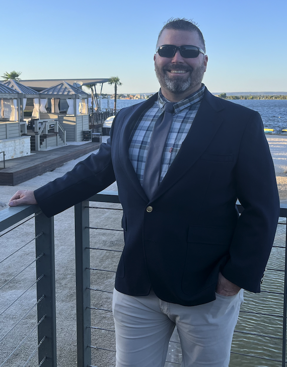 A man in sunglasses, a navy blazer, checked shirt, and tie, standing outdoors by a waterfront at sunset, smiling.