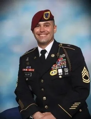 A smiling soldier in dress uniform with medals and patches, wearing a maroon beret, against a blue background.