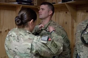A female soldier helping a male soldier adjust his military uniform in a wooden locker room.
