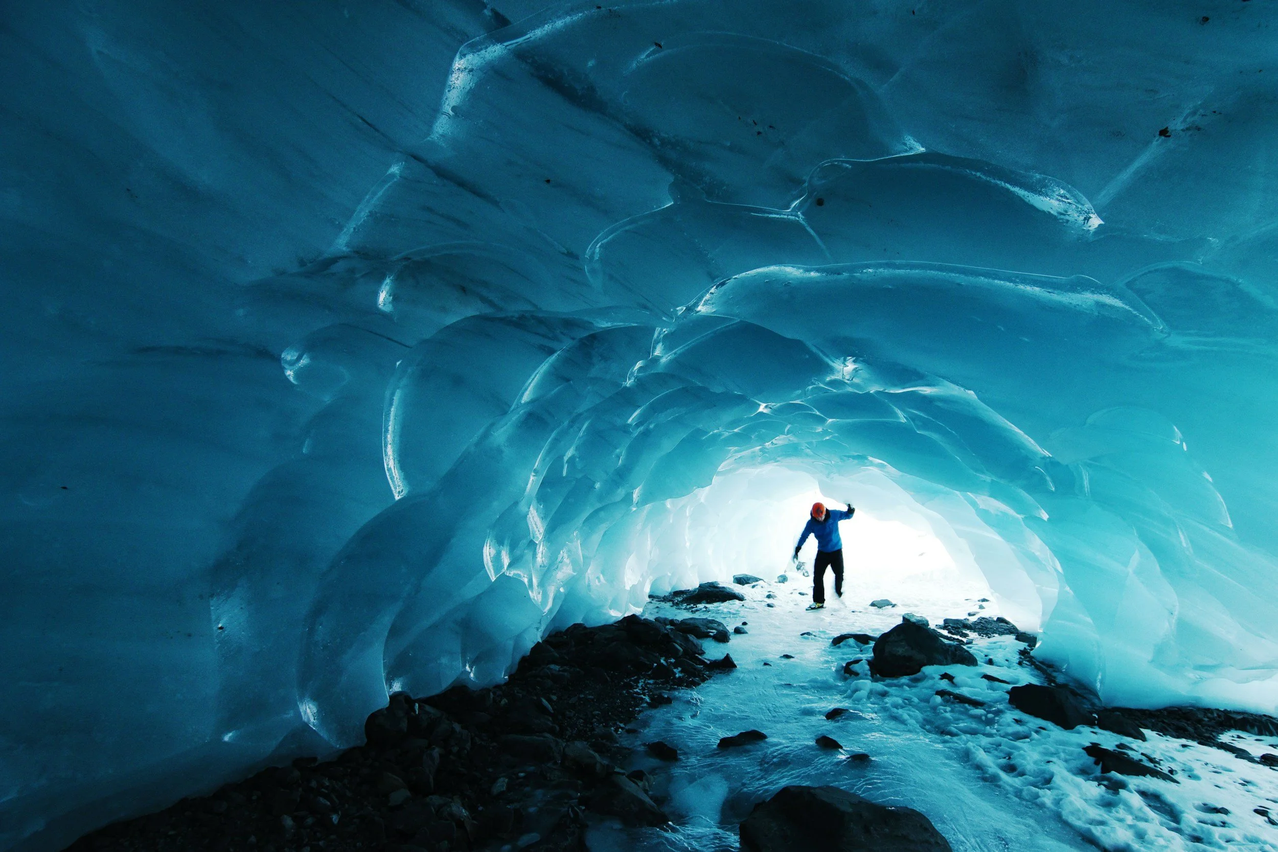 A person standing inside an ice cave with blue icy walls and ceiling, illuminated by natural light from the cave's opening.