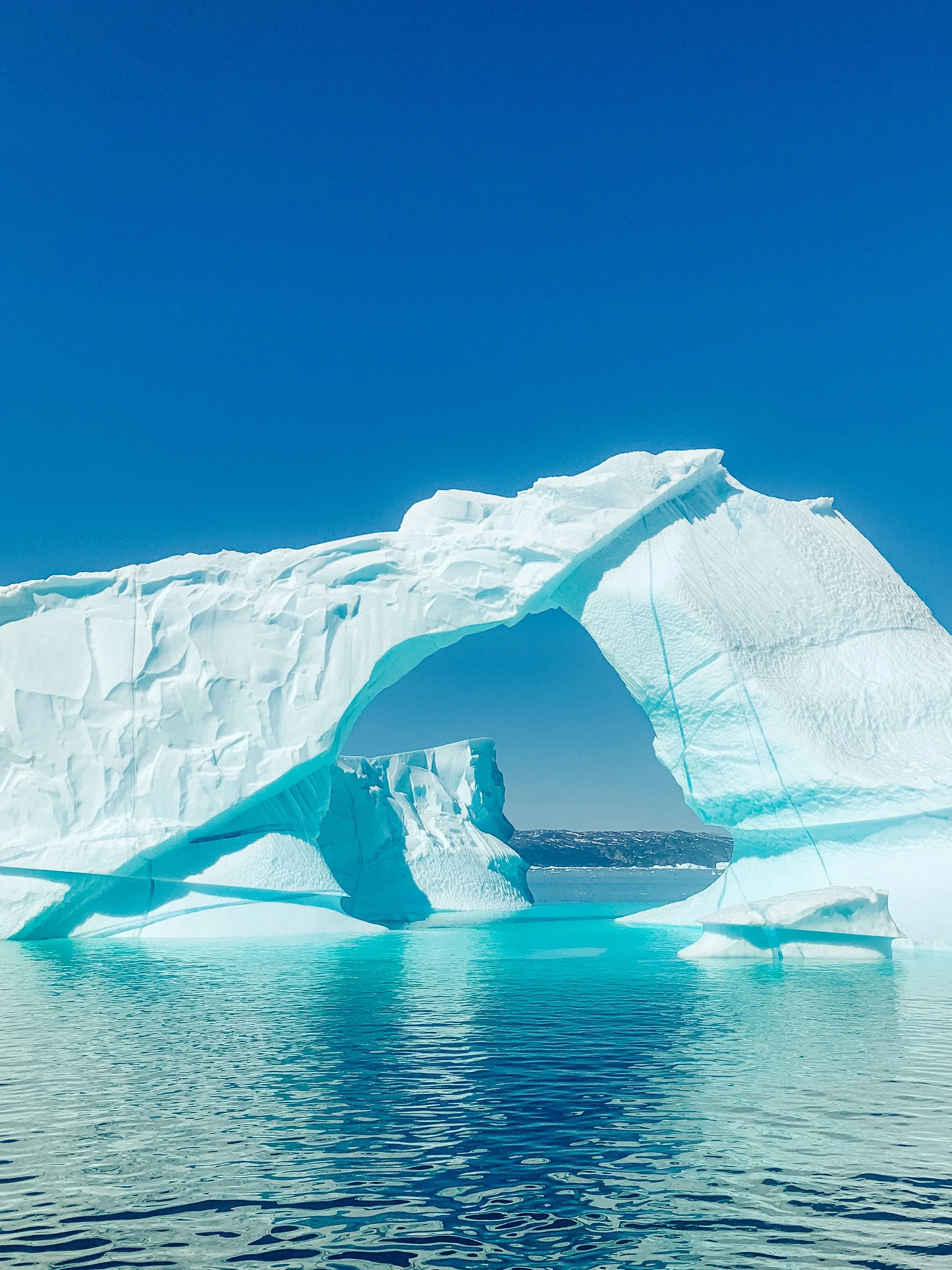 A large iceberg with a natural arch formation floating in cold ocean water under clear blue sky.