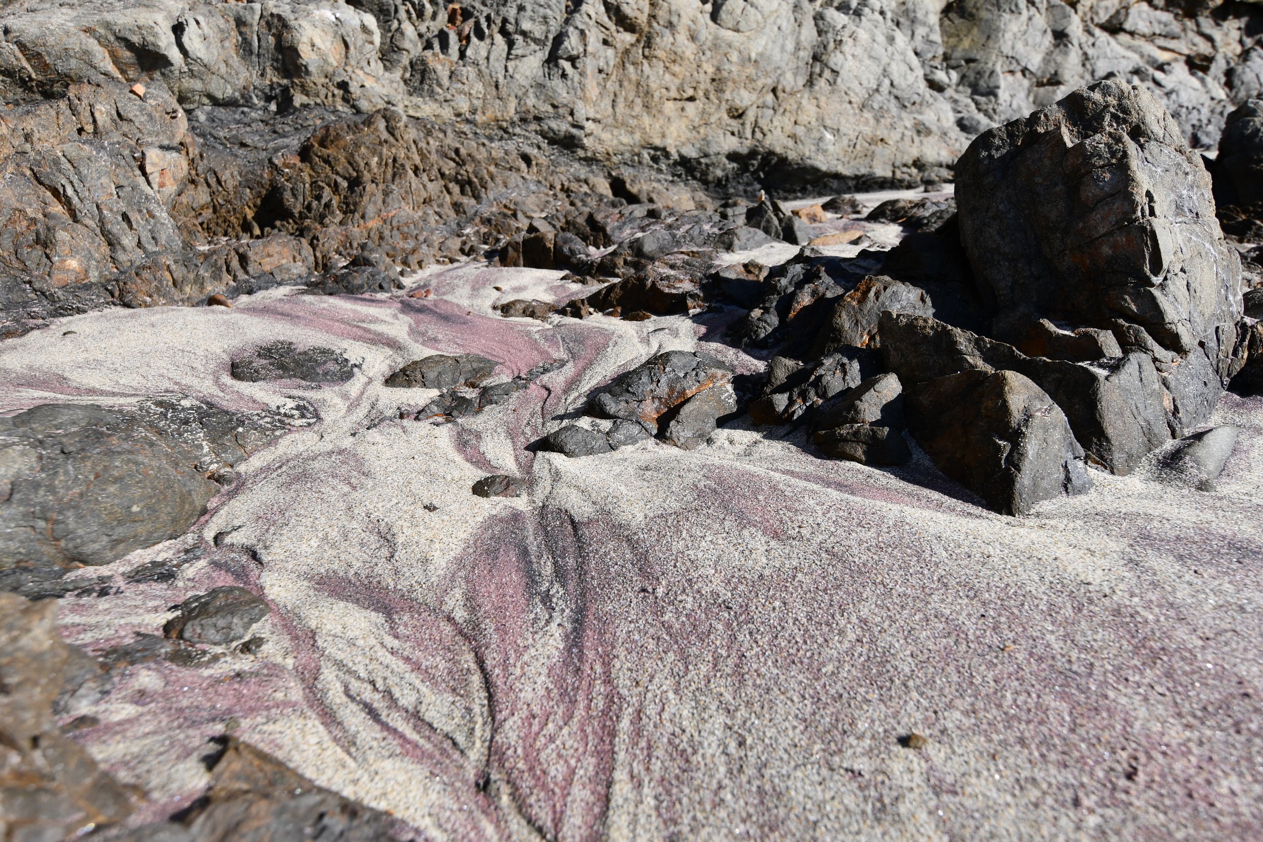 Felsiger Strand mit bunten Sedimentlagen und schwarzen Steinen in einer felsigen Küstenlandschaft.
