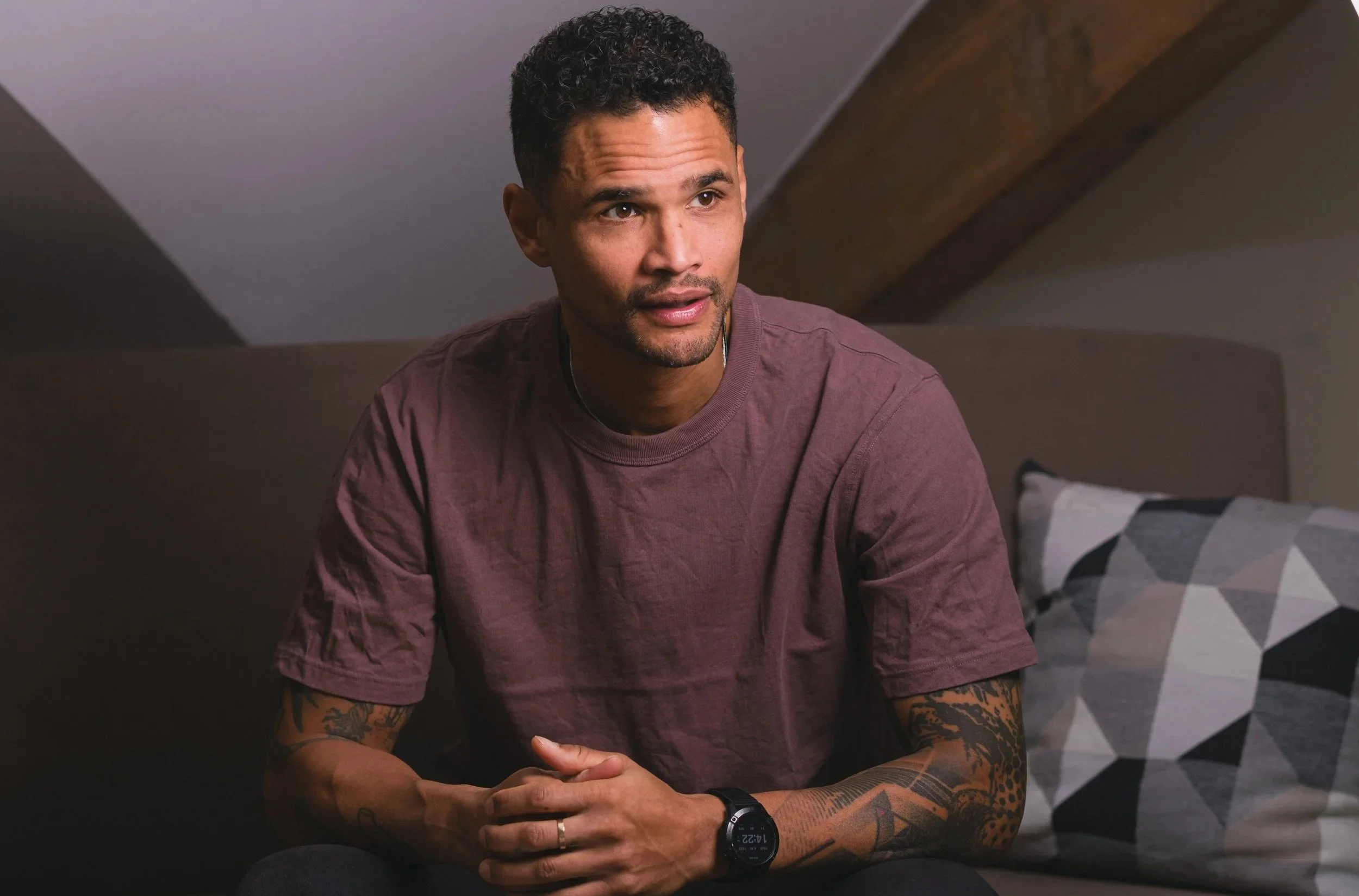 Junger Mann mit Tattoos sitzt auf einem Sofa, trägt einen dunklen T-Shirt und eine Smartwatch, spricht und hört aufmerksam.