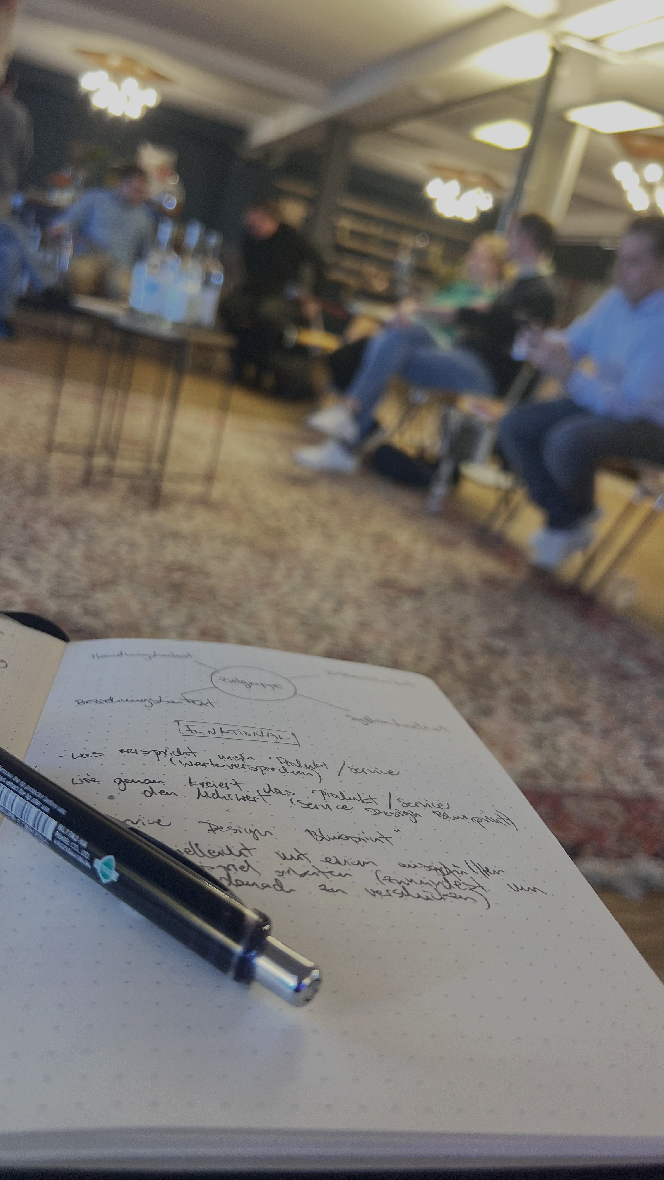 Blick auf ein Notizbuch mit Kugelschreiber im Vordergrund, während im Hintergrund eine Gruppe von Menschen in einem Raum bei einem Treffen sitzt.