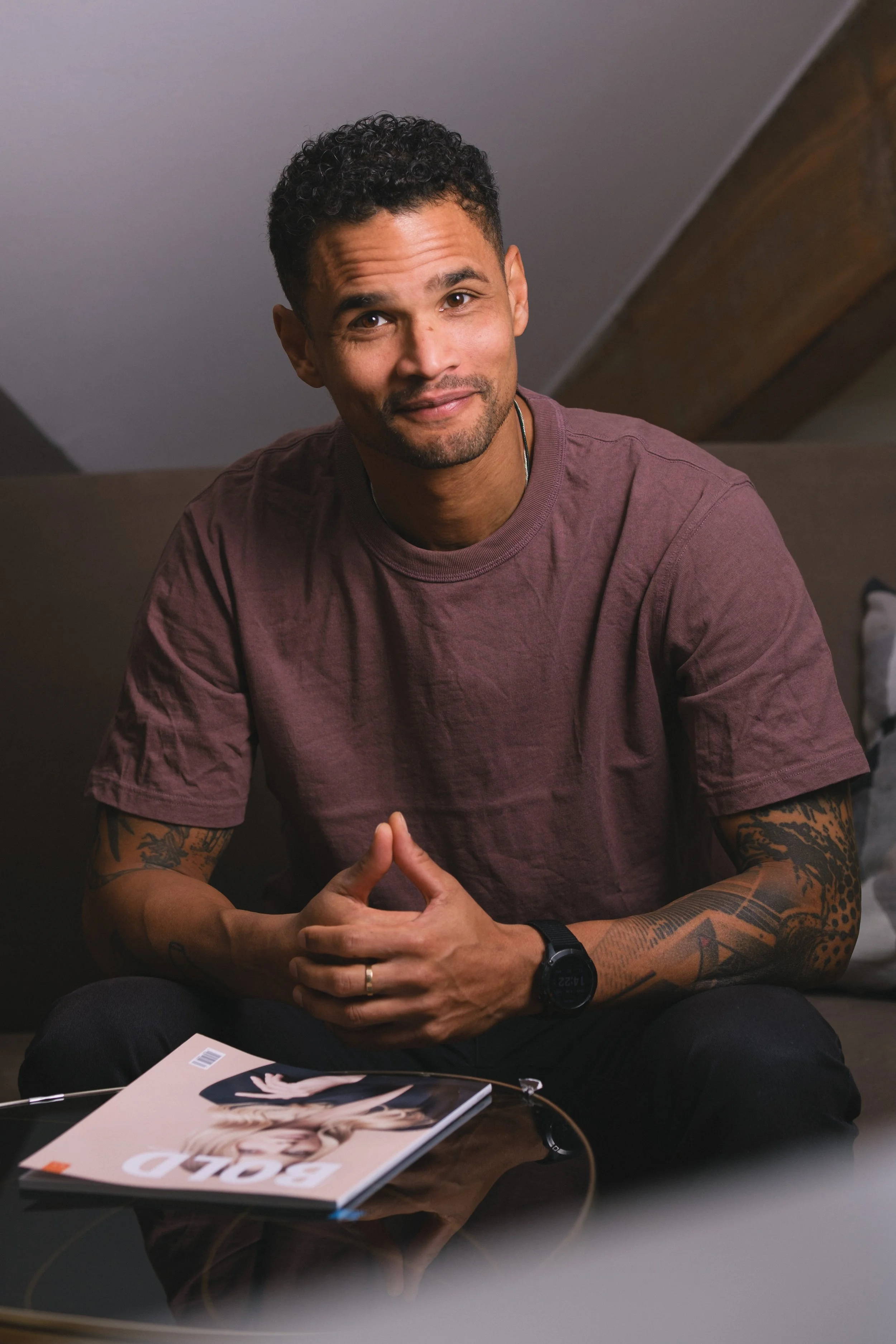 Ein Mann mit dunklen, curly Haaren, Tattoos auf den Armen, trägt eine Armbanduhr und sitzt an einem Tisch mit einer Zeitschrift.