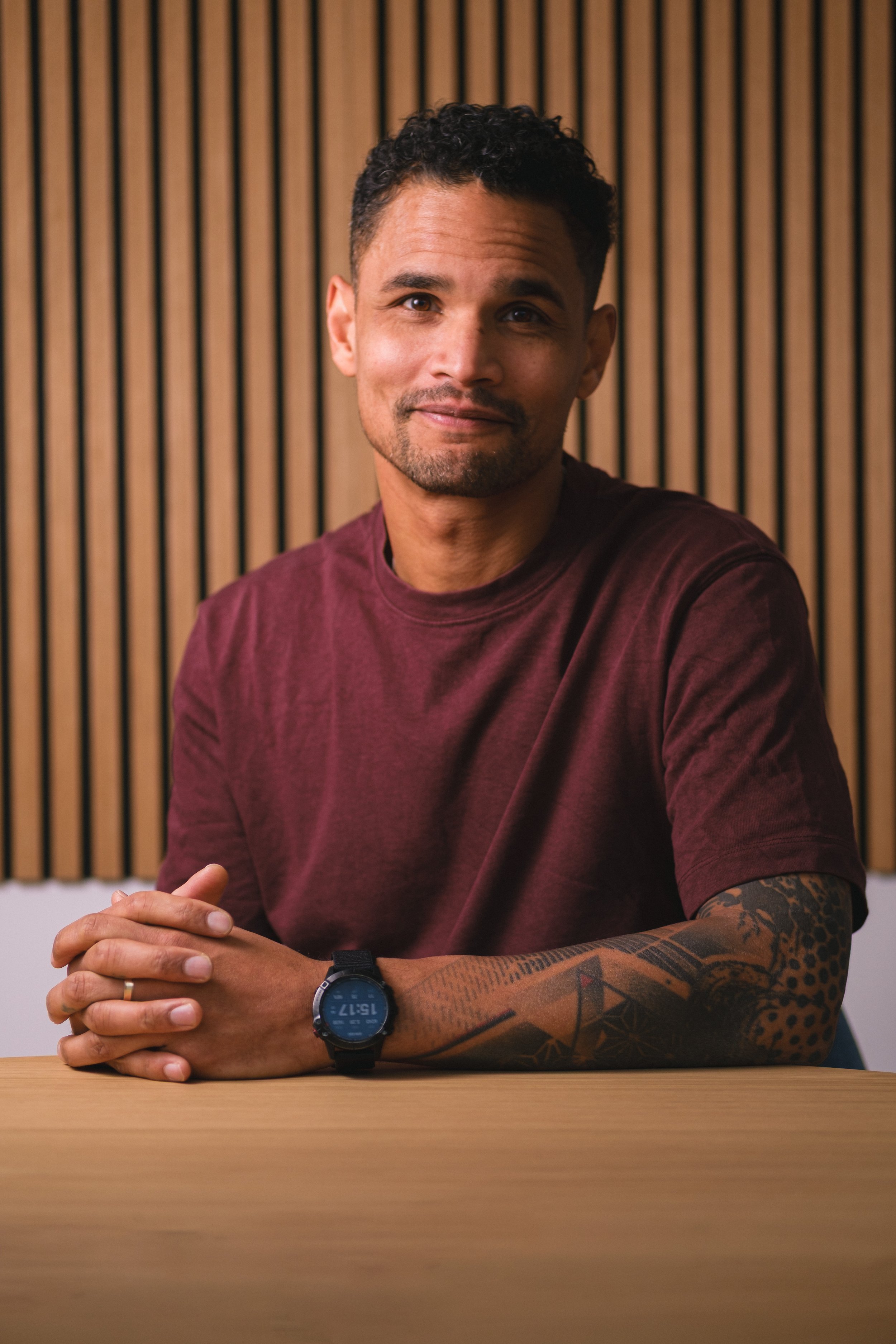 Ein Mann sitzt an einem Tisch vor einer Holzvertäfelung, trägt ein maroonfarbenes T-Shirt und eine Armbanduhr, zeigt ein freundliches Lächeln und hat Tattoos auf seinem linken Arm.