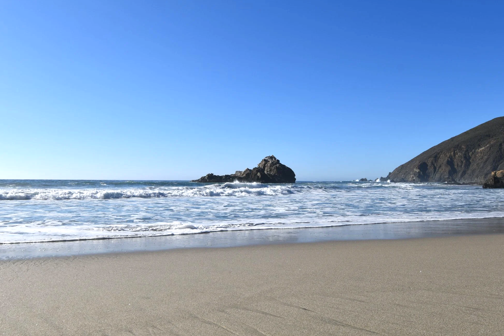 Strand mit Sand, in der Mitte Felsen im Meer, auf der rechten Seite eine Klippe, blauer Himmel.