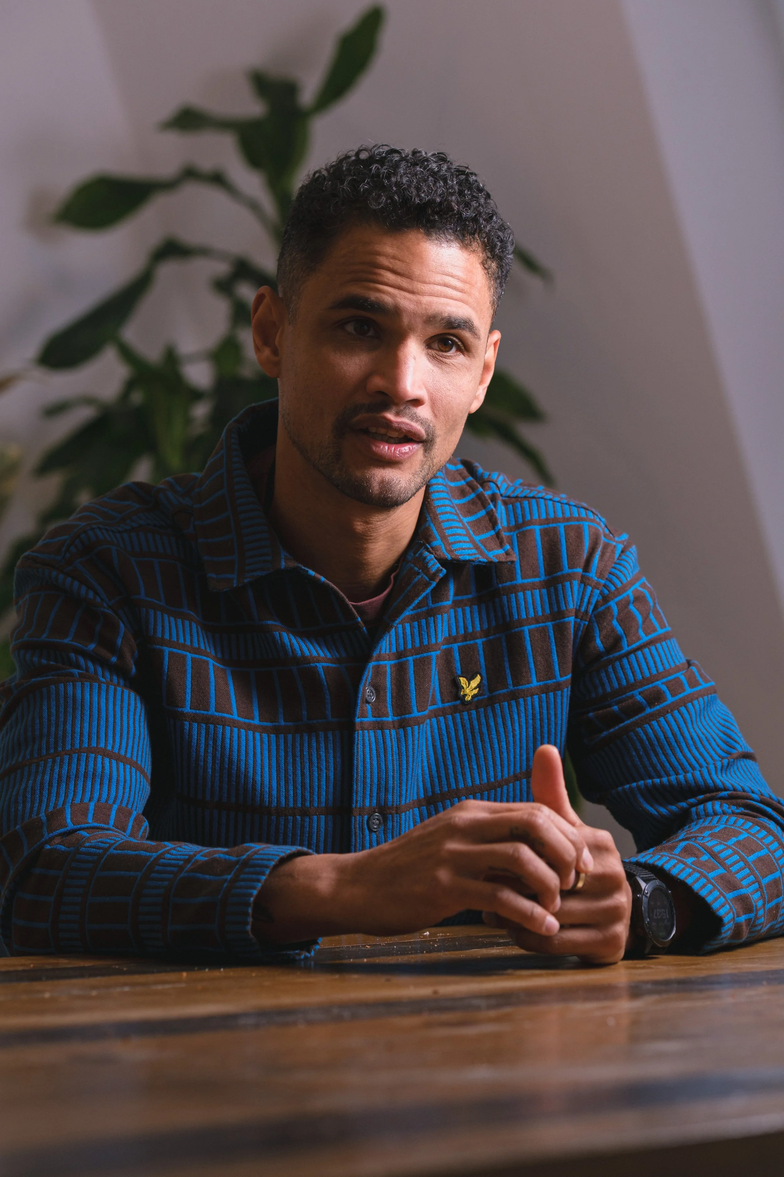 Ein Mann sitzt an einem Holztisch und spricht, im Hintergrund eine Pflanze und eine helle Wand.